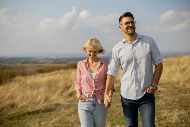 Parlak bir gökyüzünün altında çimenli bir yamaçta el ele yürüyen iki kişi. Gülüyorlar ve güneşli bir öğleden sonra birlikte doğada vakit geçiriyorlar..