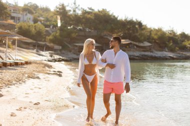 Güneş batarken, sıcak ve sevgi saçan bir çift, sakin bir plajda el ele yürür. Gülüşleri anın neşesini yansıtıyor..