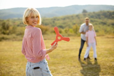 Bir kadın elinde parlak kırmızı bir oyuncak tutarken gülümsüyor, çimen tarlasında duruyor. Arka planda, bir adam ve bir çocuk güneşli, yeşil bir arazide birlikte eğleniyorlar..
