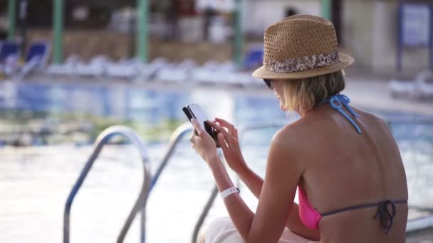 Mujer en traje de baño sentada junto a la piscina surfeando en el teléfono inteligente — Vídeo de stock