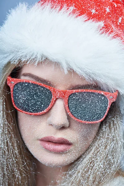 Chica congelada con nieve en la cara con sombrero de Santa y gafas de sol —  Fotos de Stock