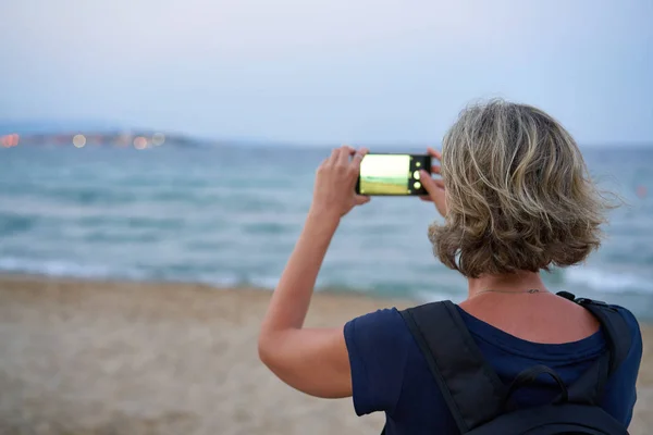 Woman taking a photo of a sea on smart phone on sunset — ストック写真