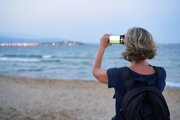 Woman taking a photo of a sea on smart phone on sunset — ストック写真