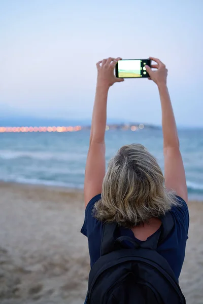 Woman taking a photo of a sea on smart phone on sunset — ストック写真