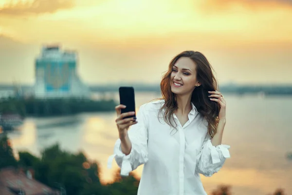 Woman making self portrait with her smart phone over cityscape golden sunset — ストック写真