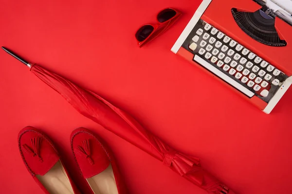 Φωτεινά κόκκινα παπούτσια, ομπρέλα, γραφομηχανή και γυαλιά που βρίσκονται σε κόκκινο φόντο — Φωτογραφία Αρχείου