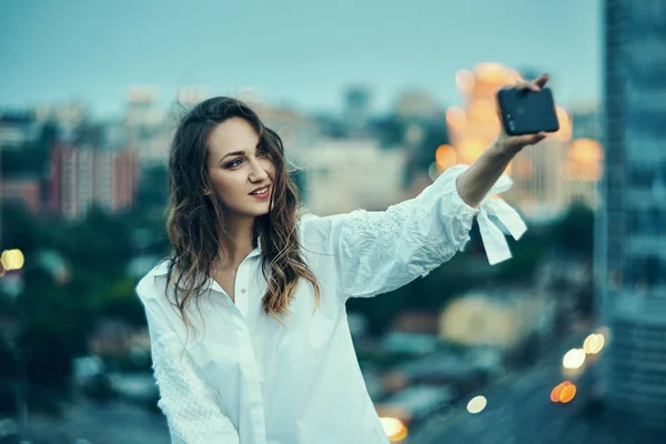 Mujer haciendo autorretrato con su teléfono inteligente sobre la puesta de sol de oro paisaje urbano — Foto de Stock