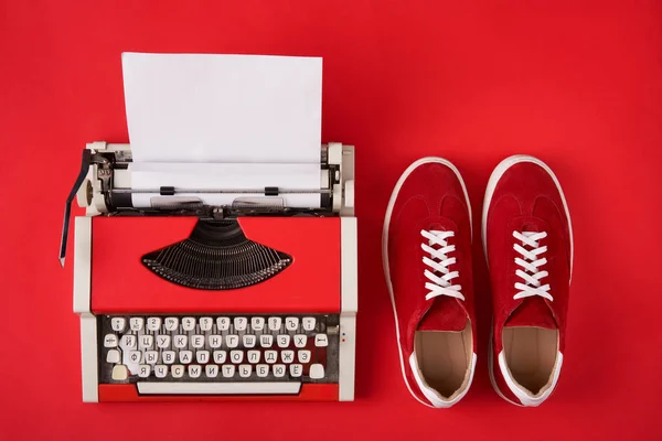 Sapatilhas de camurça vermelha e máquina de escrever com folha de papel em branco no fundo vermelho — Fotografia de Stock