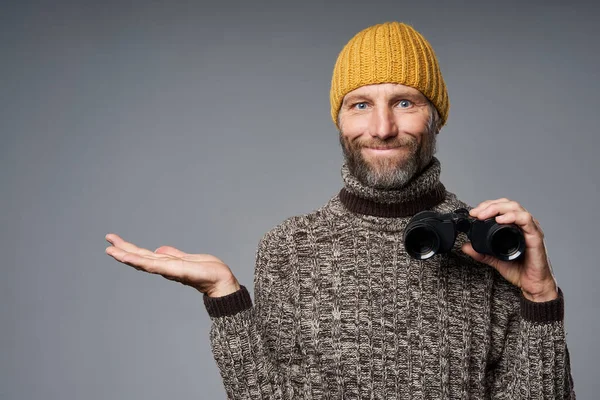 Сосредоточенный взрослый мужчина в тёплом свитере и вязаной шляпе, держащий бинокль — стоковое фото