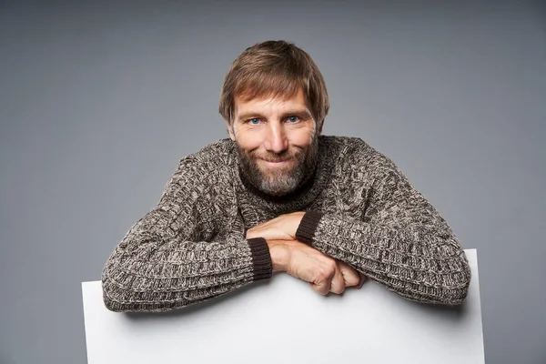 Uśmiechnięty dojrzały mężczyzna w ciepłym swetrze opierający się na białym transparencie — Zdjęcie stockowe