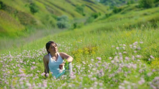 Молода жінка сидить на зеленому лузі і диких квітах насолоджується весняним літнім сонячним світлом — стокове відео