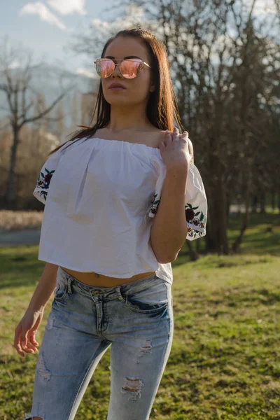 美丽的年轻女子穿着时髦的牛仔裤和白色上衣 在夏季公园里摆着花饰 — 图库照片