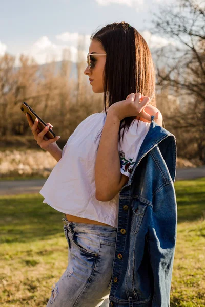 attractive woman in sun glasses and casual clothes using smartphone while walking on meadow