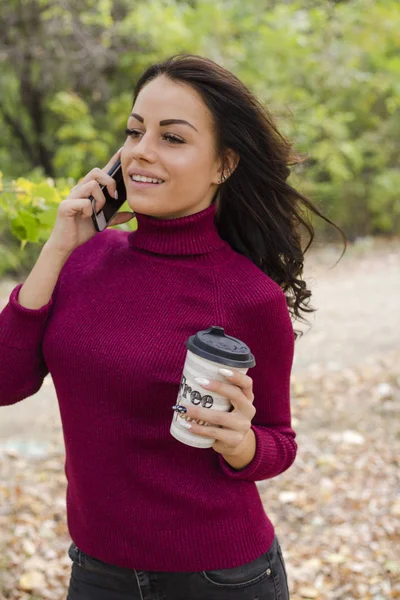 portrait of beautiful young woman in sweater with paper cup of coffee talking by phone in park