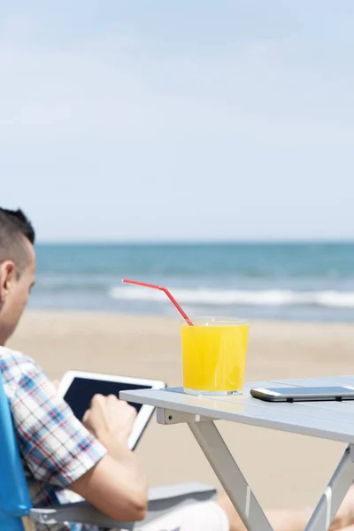 特写在海滩上的一个年轻的白种人 使用数字平板电脑 坐在户外桌子上 一杯清爽的橙饮料和智能手机上 — 图库照片