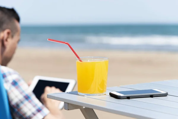 特写在海滩上的一个年轻的白种人 使用数字平板电脑 坐在户外桌子上 一杯清爽的橙饮料和智能手机上 — 图库照片