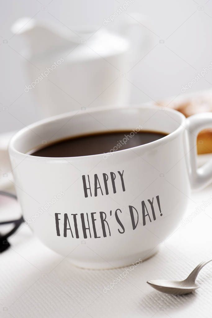 closeup of a white ceramic cup with coffee, with the text happy fathers day written in it, at a table set for breakfast