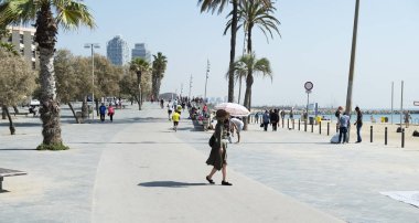 BARCELONA, SPAIN - APRIL 24, 2018: People walking and running by the seafront of the popular La Barceloneta beach in Barcelona, Spain, with the Mapfre Tower and the Hotel Arts in the background clipart