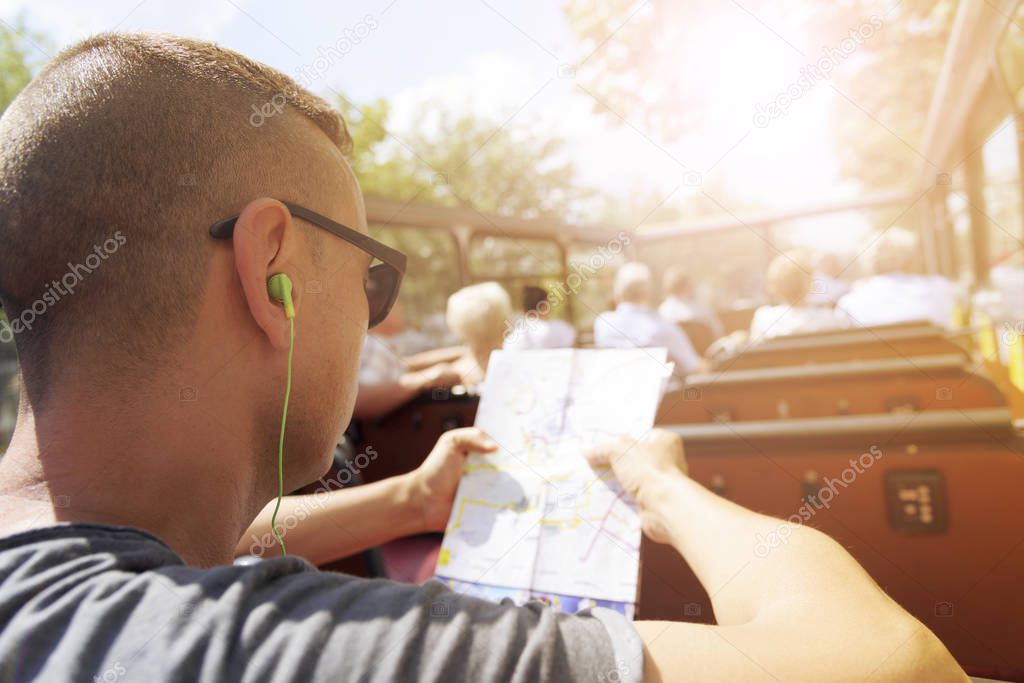 closeup of a young cacausian man traveling on an open top tour bus, checking a city map and wearing earphones, with a sunbeam in the background
