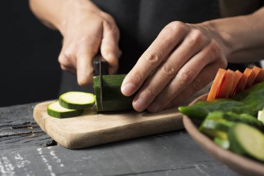 bir kabak havuç veya yeşil biber gibi bazı farklı sebzeli tepsi yanında rustik ahşap bir masa üzerinde bir bıçakla dilimler halinde keserek bir siyah önlük giyen bir beyaz adam, closeup