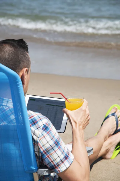 一个年轻的白种人在海滩上特写 使用数字平板电脑 坐在蓝色的甲板椅上 手里拿着一杯清爽的橙汁饮料 — 图库照片