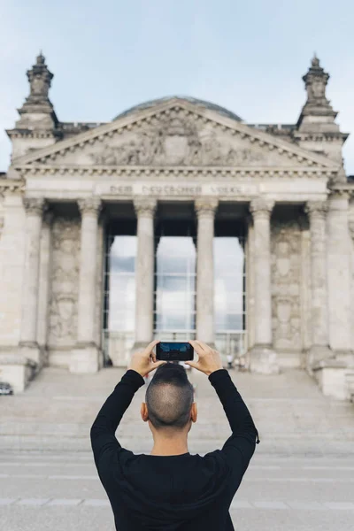 特写一个年轻的白种人 从后面看到 在德国柏林的国会大厦门面的照片 与他的智能手机 — 图库照片