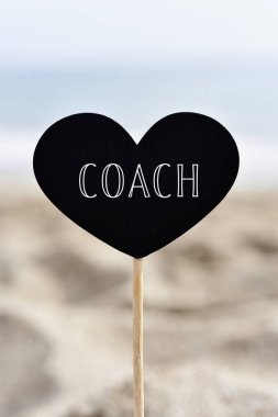 closeup of a black heart-shaped signboard with text coach written in it, attached to a stick, on the sand of a beach, with the sea in the background clipart