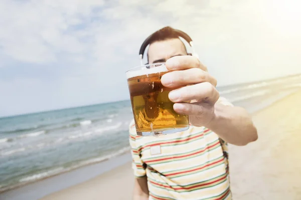 浜辺の中無線ヘッドフォンで音楽を聴く若い白人男のクローズ アップはバック グラウンドで太陽光線が付いている海の前でビールを飲みながら — ストック写真