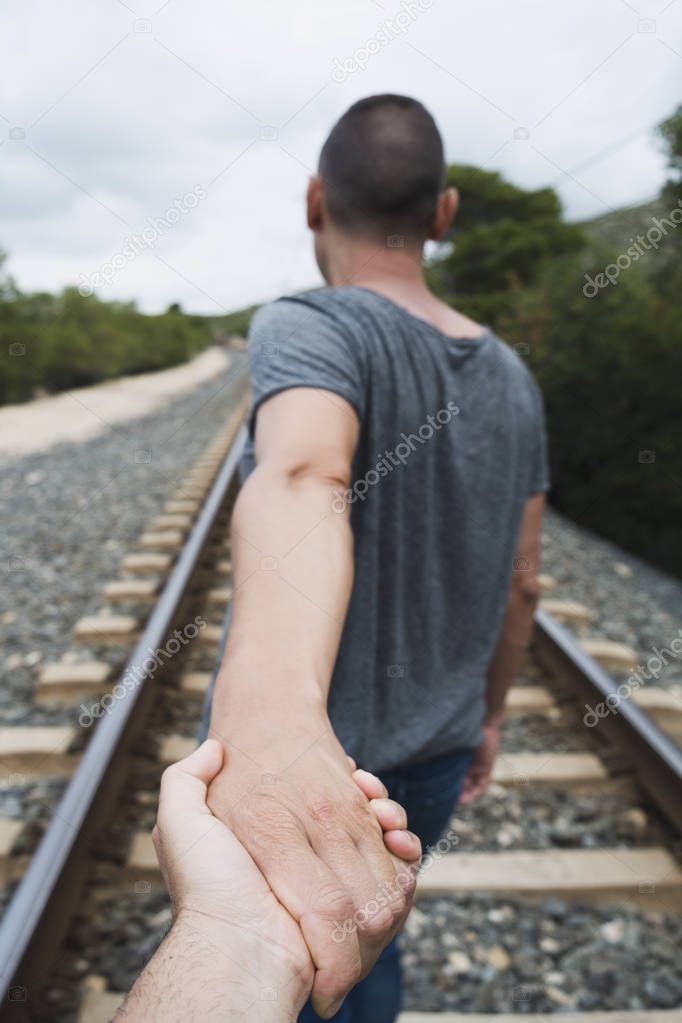 closeup of the hand of a caucasian man holding the hand of another caucasian man, seen from behind, walking by the railroad tracks on a natural landscape