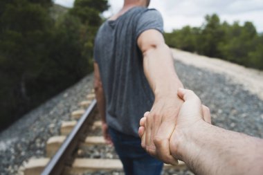 closeup of the hand of a caucasian man holding the hand of another caucasian man, seen from behind, walking by the railroad tracks on a natural landscape clipart