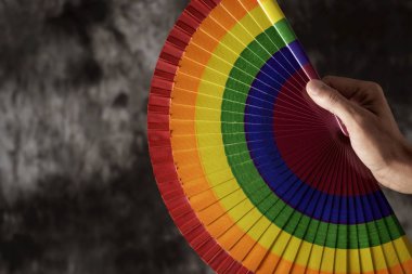 closeup of the hand of a young caucasian man with a rainbow-patterned hand fan against a dark background with some blank space on the left clipart