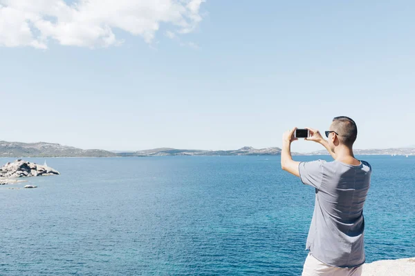 从后面看到一个年轻的高加索男子特写拍摄在哥斯达黎加 Smeralda 在意大利撒丁岛的海的照片 与他的智能手机 在上面的空白空间 — 图库照片