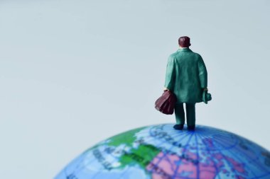 miniature traveler man seen from behind carrying a suitcase, on the top of the terrestrial globe against an off-white background with some blank space clipart