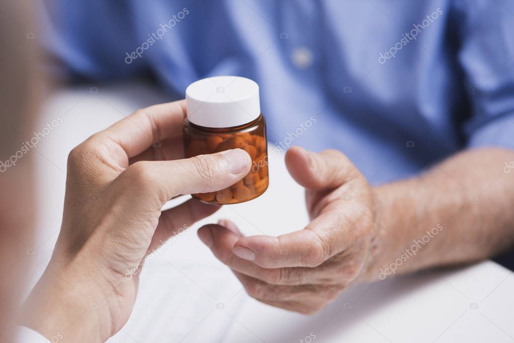closeup of a caucasian doctor man, in a white coat, giving a bottle of pills to a senior caucasian patient man, sitting both at a doctors desk