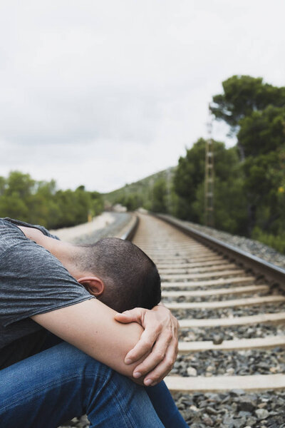 a caucasian man, wearing casual clothes, curled up on the railroad tracks, in a natural landscape