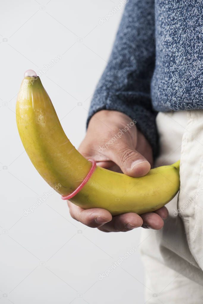 closeup of the a young caucasian man holding a banana with a pink condom in it in front of his crotch