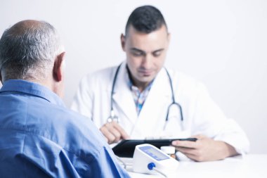 closeup of a senior caucasian patient man seen from behind and a caucasian doctor man checking his medical history or the results of a test in his tablet, sitting both at the doctors desk clipart