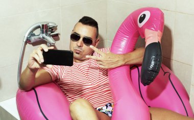 Genç bir beyaz adam, güneş gözlüğü takıyor bir yüzme takım elbise ve yüzmeye onun smartphone ile kendi portresi alarak onun banyo küvet içinde pembe flamingo şeklinde ring