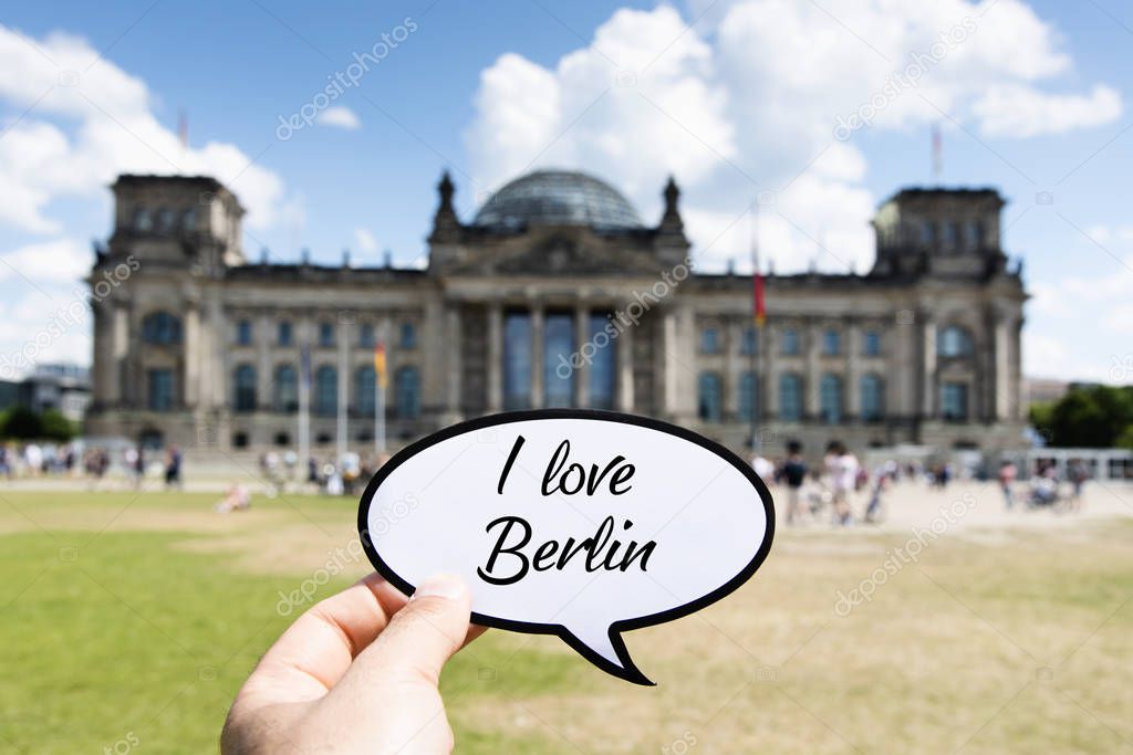 closeup of the hand of a young caucasian man showing a speech bubble with the text I love Berlin written in it, in front of the Reichstag building in Berlin, Germany