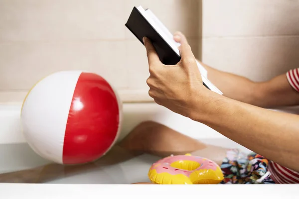 一个身穿泳装的年轻白种男人在浴室浴缸里看书 旁边是充气粉色甜甜圈和漂浮在水中的充气球 — 图库照片