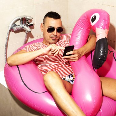 Genç bir beyaz adam, güneş gözlüğü takıyor bir yüzme takım elbise ve yüzmeye onun banyo küvetinde onun smartphone kullanarak bir pembe flamingo şeklinde ring