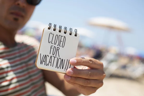 特写一个年轻的白人男子戴太阳镜在海滩上显示一个螺旋记事本与文本关闭的假期手写在第一页 — 图库照片