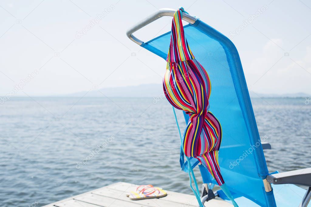 a colorful bikini drying on a blue deck chair and a pair of flip-flops on a wooden pier next to the water