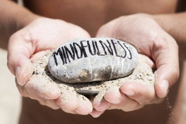 closeup of a young caucasian man, on the beach, with a handful of sand in his hands and a stone on top with the text mindfulness written in it clipart