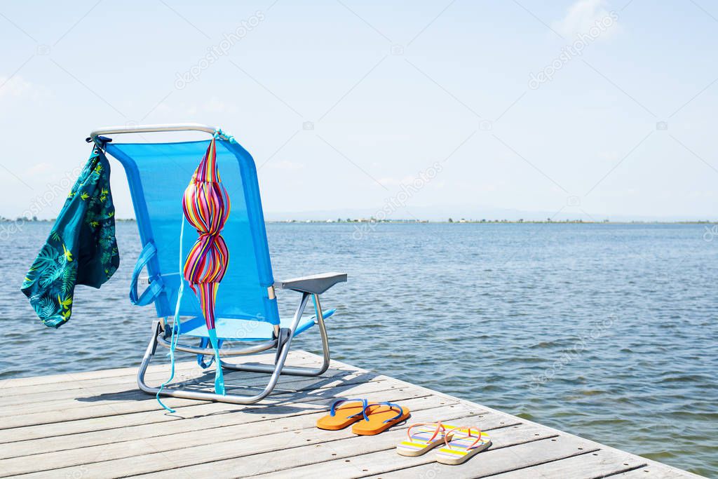a colorful bikini and a pair of blue swimming trunks drying on a blue deck chair, and two pair of flip-flops on a wooden pier, next to the water