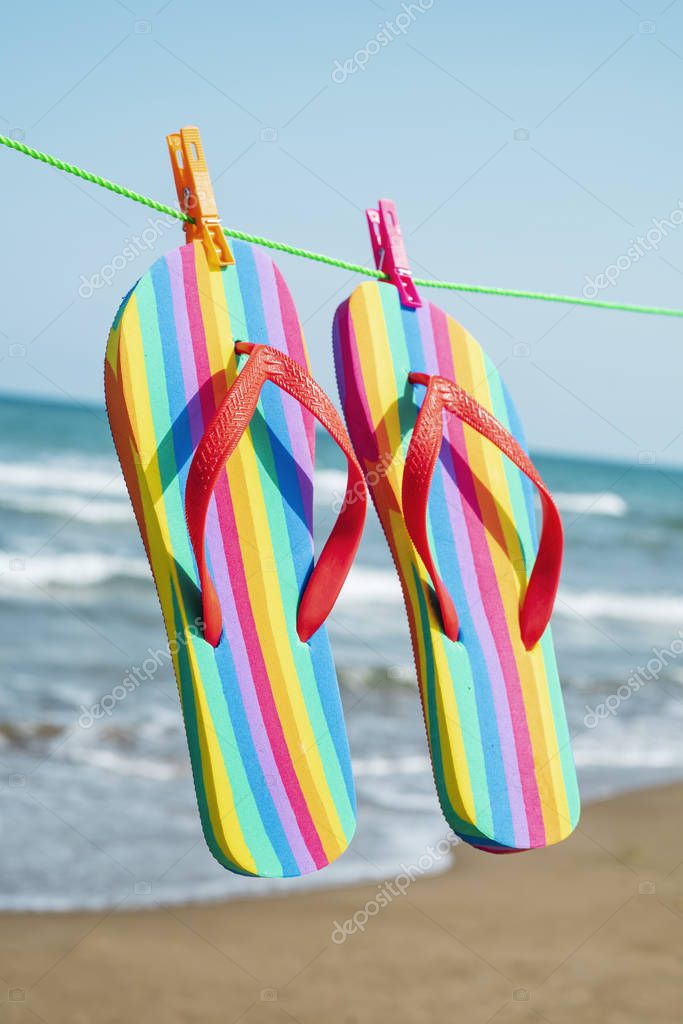 closeup of a pairs of colorful flip-flops, patterned with the colors of the rainbow flag, hanging on a clothes line on the beach, with the sea in te background