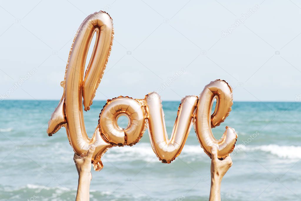 closeup of the hands of a young caucasian man on the beach holding a balloon in the shape of the word love against the sky, with the sea in the background