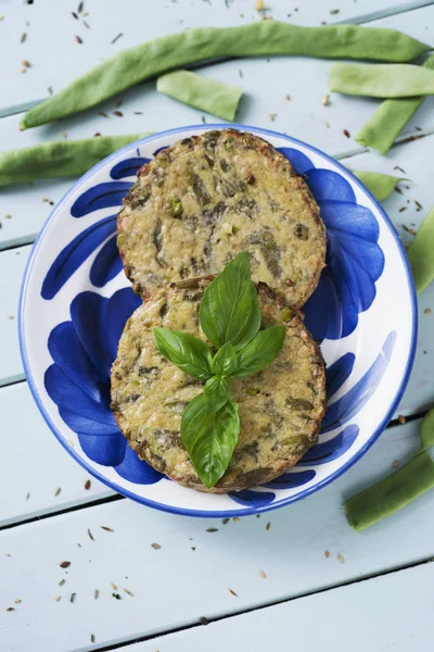 high angle view of some veggie burgers made with green beans and other vegetables in a white and blue ceramic plate, placed on a pale green rustic wooden table