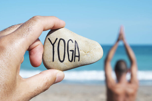 closeup of the hand of a young caucasian man on the beach holding a stone with the yoga written in it, and a young caucasian man practicing yoga in the background