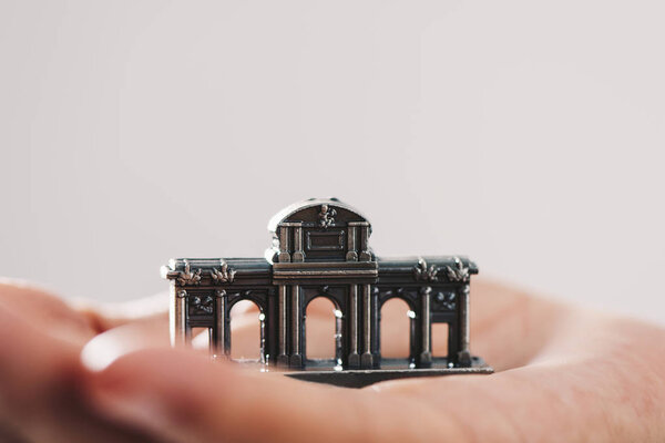a miniature of the Puerta de Alcala in Madrid, Spain, on the hand of a young man against an off-white background, with some blank space on top